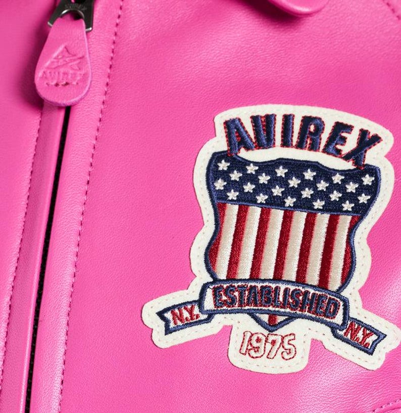 K&ouml;nnte beinhalten: Nahaufnahme einer leuchtend rosa Lederjacke. Die Jacke hat einen Aufn&auml;her mit dem Text "AVIREX" &uuml;ber einem Schilddesign mit der amerikanischen Flagge und den Worten "ESTABLISHED 1975 N.Y."