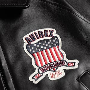 May include: Close-up of a black leather jacket featuring a patch with the word "AVIREX" in red and white lettering. The patch has a shield design with a blue field of white stars and red and white stripes. The patch also includes the text "NY ESTABLISHED 1975".
