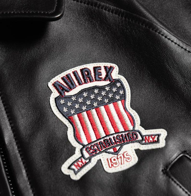 May include: Close-up of a black leather jacket featuring a patch with the word "AVIREX" in red and white lettering. The patch has a shield design with a blue field of white stars and red and white stripes. The patch also includes the text "NY ESTABLISHED 1975".