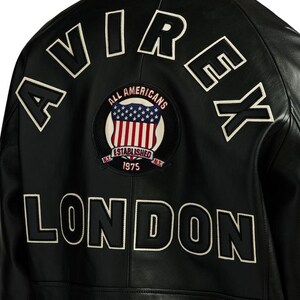 May include: Black leather jacket with white embroidered lettering spelling "AVIREX" and "LONDON". A circular patch features a US flag design with the words "ALL AMERICANS ESTABLISHED 1975".