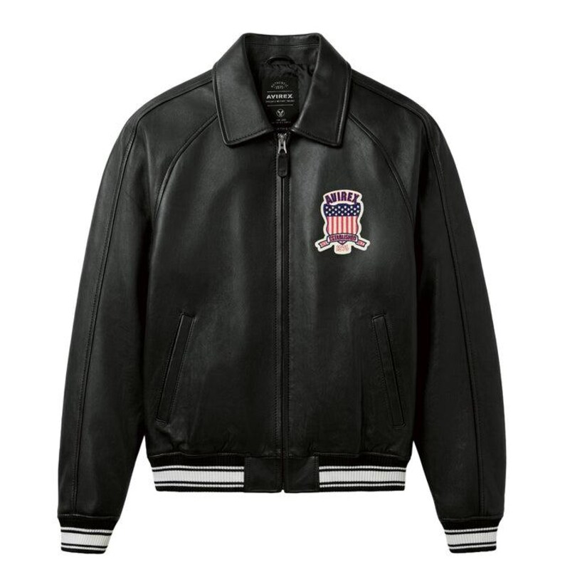 May include: Black leather jacket with a collared neckline, a zip-up front, and a patch on the chest featuring an American flag design. The jacket has white and black striped cuffs and hem. The brand name "AVIREX" is visible on the jacket.