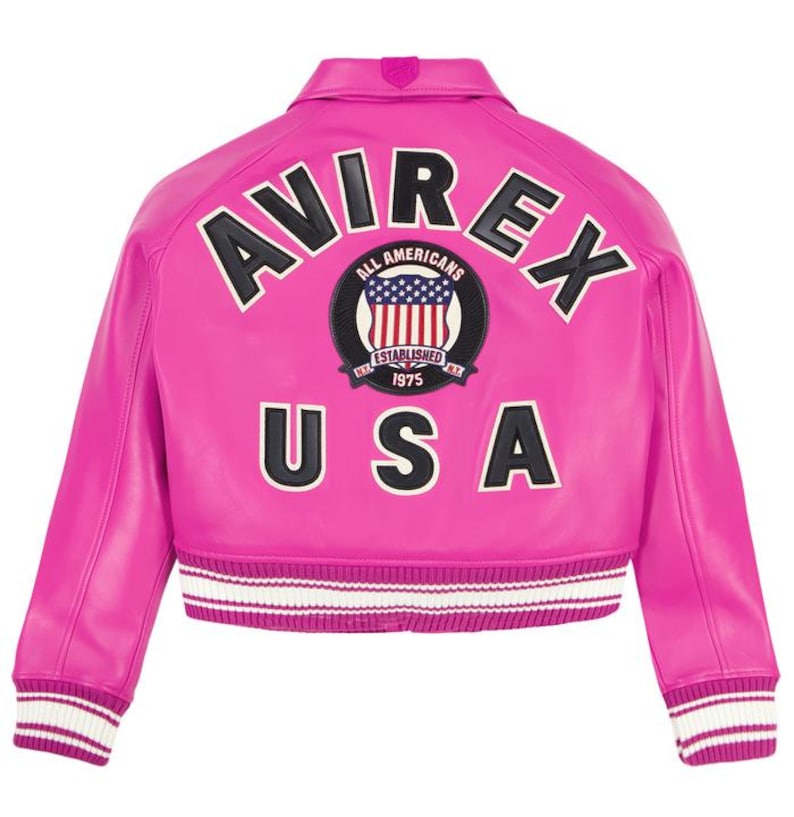 K&ouml;nnte beinhalten: Eine leuchtend pinkfarbene Lederjacke mit dem Wort "AVIREX" in schwarz-wei&szlig;en Buchstaben &uuml;ber den Schultern. Darunter befindet sich ein kreisf&ouml;rmiger Aufn&auml;her mit den Worten "ALL AMERICANS" und "ESTABLISHED 1975". Das Wort "USA" steht darunter.