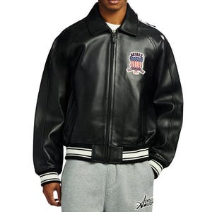 May include: Black leather jacket with a zip-up front, a collar, and a patch on the chest with an American flag design. The jacket has white stripes on the cuffs and waistband. The brand name "Aviirex" is visible on the lower right.
