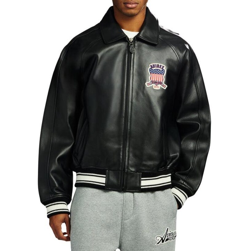 May include: Black leather jacket with a zip-up front, a collar, and a patch on the chest with an American flag design. The jacket has white stripes on the cuffs and waistband. The brand name "Aviirex" is visible on the lower right.