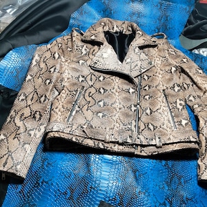Pode incluir: Uma jaqueta estilo motociclista com um padrão de pele de cobra em tons de bege e castanho. A jaqueta inclui uma gola entalhada, mangas compridas com punhos com fecho, bolsos verticais com fecho e um cinto com fivela na bainha. O interior é forrado a preto.