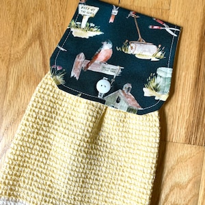 May include: A yellow and white waffle weave kitchen towel with a dark green fabric pocket. The pocket has a print of birds, gardening tools, and a sign that says "Keep off the grass".