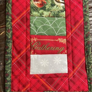 May include: A quilted Christmas table runner with a red plaid design and a green border. The center features fabric panels with birds, a green geometric pattern, the word "Gathering", and snowflakes. A festive holiday decoration.