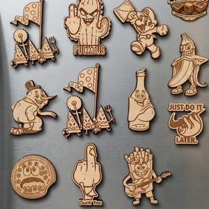 May include: A collection of laser-cut wooden magnets featuring various cartoon characters and objects, including a cactus, a running figure, a banana, a snowman, a bottle, a cookie, a duck, and french fries, with text labels like FUCKTUS and DUCK YOU.