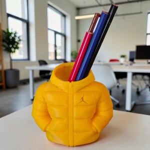 Peut inclure: Un porte-crayons jaune en forme de doudoune, avec un petit logo sur le devant. Il contient des crayons rouges, bleus, marron et noirs. Le porte-crayons est posé sur une surface blanche.