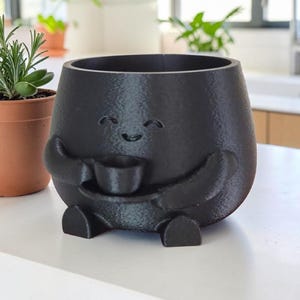 Peut inclure: Un pot de fleurs noir souriant avec des bras et des jambes, tenant une petite tasse. Le pot a une surface texturée et un design fantaisiste. Une petite plante en pot est visible en arrière-plan.