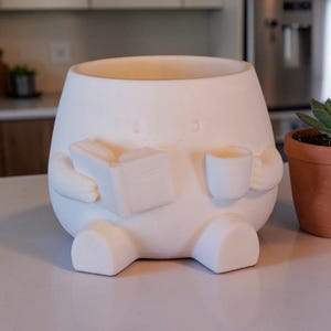 Op de afbeelding: Een witte keramische plantenpot in de vorm van een stripfiguur, met een boek en een kopje. De pot heeft een rond lichaam, korte benen en armen. Rechts staat een kleine terracotta pot met een vetplant.