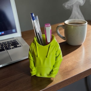 Peut inclure: Un organiseur de bureau en forme de sweat à capuche vert citron contient des stylos et une gomme rose et blanche. Un ordinateur portable et une tasse de café fumant sont en arrière-plan. L'organisateur est sur un bureau en bois.
