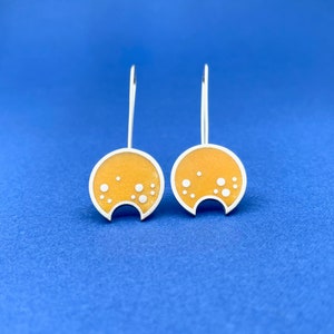 May include: A pair of silver dangle earrings with a yellow enamel disc featuring white dots. The discs have a semi-circular cut out at the bottom.