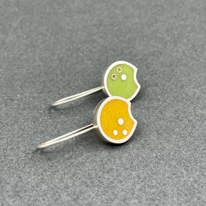 May include: A pair of silver earrings with a unique design. Each earring features a rounded, crescent-shaped charm in either yellow or green, with white dots and a silver border. The earrings have a simple, curved hook.