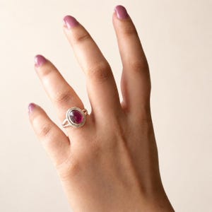 May include: A silver ring featuring a large, oval, deep pink gemstone. The ring has a textured, rope-like border around the stone. The ring is on a hand with painted nails.