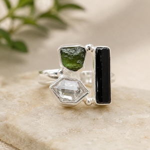 Peut inclure: Bague en argent ornée de trois pierres : une pierre verte rectangulaire, une pierre transparente taillée géométriquement et une pierre noire rectangulaire. La bague est posée sur une surface claire et texturée.