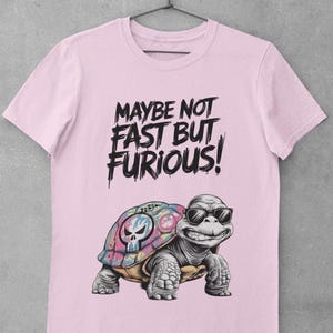 May include: Light pink t-shirt featuring a smiling turtle wearing sunglasses. The turtle has a colorful shell with a skull design. The shirt has the text "MAYBE NOT FAST BUT FURIOUS!" in black, stylized font.