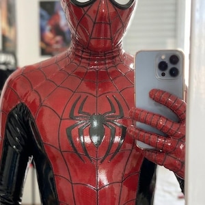 Puede incluir: Un disfraz de Spider-Man de cuerpo completo en rojo y negro con acabado brillante, un emblema de ara&ntilde;a negro en el pecho y un patr&oacute;n de telara&ntilde;a en las secciones rojas. El traje incluye una m&aacute;scara a juego con lentes de malla blanca y es sostenido por un smartphone azul claro.