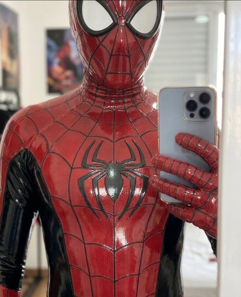 Puede incluir: Un disfraz de Spider-Man de cuerpo completo en rojo y negro con acabado brillante, un emblema de ara&ntilde;a negro en el pecho y un patr&oacute;n de telara&ntilde;a en las secciones rojas. El traje incluye una m&aacute;scara a juego con lentes de malla blanca y es sostenido por un smartphone azul claro.