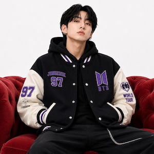 May include: A black and cream varsity jacket featuring purple embroidery with the text JUNGKOOK 97, the BTS logo, and a heart graphic with the text WORLD TOUR. The jacket has a hood, snap button closures, and striped ribbed cuffs and collar.