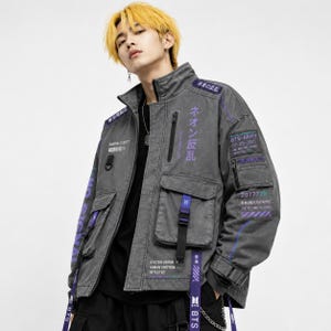Puede incluir: Chaqueta vaquera gris con múltiples bolsillos y detalles morados. Incluye texto en japonés e inglés, como "BTS-ARMY" y "207777". La chaqueta tiene cuello alto, cremallera y correas con hebillas.