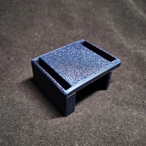 May include: A small, rectangular, dark blue 3D-printed stand with a glittery, textured top surface. The object features a raised platform design with open sides and a solid base, showcasing visible layer lines from the manufacturing process.