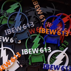 May include: A collection of colorful 3D-printed logos featuring the text "IBEW 613" and a lightning bolt design. The logos are in various colors, including white, orange, blue, green, and red, against a dark background.