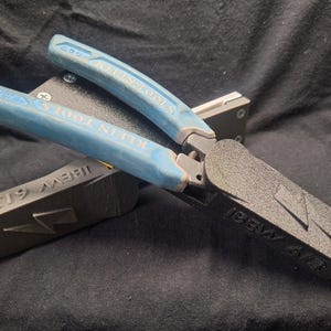 May include: Klein Tools pliers with light blue rubberized handles featuring a black 3D-printed attachment on the jaws. The attachment is embossed with the text IBEW 613 and a lightning bolt symbol. The pliers are resting on a matching black 3D-printed block.