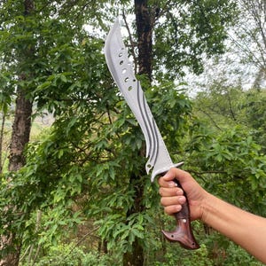 May include: A curved silver steel blade featuring a series of circular cutouts along the spine and decorative wavy grooves on the flat surface. The handle is crafted from polished dark brown wood with a distinct hooked pommel and metal rivets.