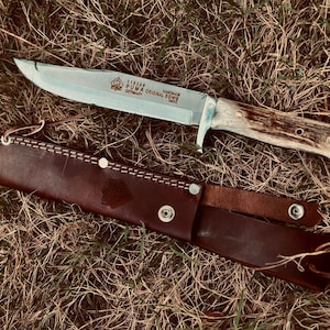 May include: A Puma Original Bowie knife with a silver steel blade featuring engraved text and a textured antler handle, accompanied by a dark brown leather sheath with silver metal snap buttons and a stamped logo on the front surface.