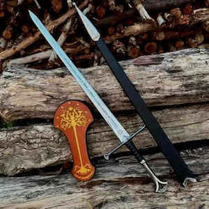 Può includere: Una lunga spada in metallo con lama appuntita, guardia trasversale e impugnatura nera, accompagnata da un fodero nero con punta argentata e una placca arancione-marrone a forma di scudo con un albero dorato e una corona.