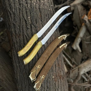 May include: Two curved knives with light yellow marbled handles and silver blades featuring intricate etched patterns, accompanied by two brown suede sheaths with polished gold-tone metal accents at the tips and throat.
