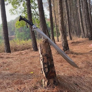 Pode incluir: Uma espada longa e curva com uma lâmina de metal prateado e um cabo preto com um pomo distinto. A espada está cravada no topo de um toco de madeira vertical e desgastado, exibindo uma ponta afiada e um design de gume curvo.