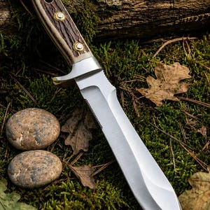 Pode incluir: Uma faca de lâmina fixa com uma lâmina de aço prateado com uma secção serrilhada no dorso e um cabo castanho texturado com três rebites de latão. A faca tem um reforço de metal que separa a lâmina do cabo com extremidade curva.