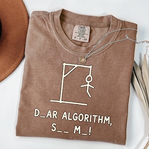 May include: A brown short-sleeved t-shirt featuring a white graphic of a stick figure on a gallows and the text "D_AR ALGORITHM, S__ M_!". The shirt has a crew neckline and a Comfort Colors brand label printed on the inside of the collar.