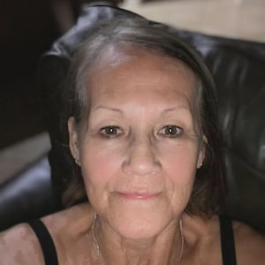 May include: A woman with fair skin and graying hair, wearing a black tank top and a silver necklace. The image is a close-up, focusing on her face and upper body. The background is blurred, suggesting an indoor setting.