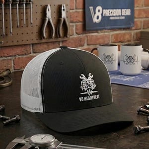 Puede incluir: Gorra de camionero negra y blanca con el logotipo V8 Heartbeat. La gorra tiene la parte trasera de malla y una visera curva. La imagen también muestra herramientas, tazas y un plano.