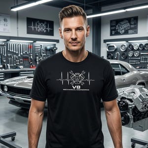 Puede incluir: Camiseta negra con un gráfico blanco de un motor V8, llaves inglesas y una línea de latidos, con el texto "V8 MECHANICAL ENGINEERING". La imagen está ambientada en un garaje con herramientas y un coche clásico.