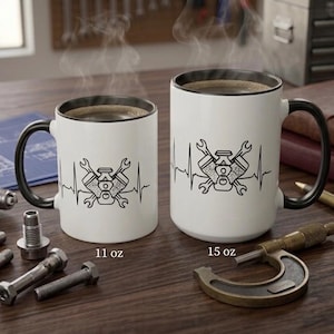 May include: Two white coffee mugs with black rims and handles, each with a black graphic of an engine and tools. The mugs are labeled 11 oz and 15 oz. Various mechanic tools and parts are scattered around the mugs on a wooden surface.