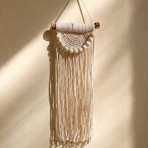 May include: A wall hanging made of off-white cotton cord knotted in a semi-circular pattern, featuring a row of smooth wooden beads along the bottom edge. The cord is suspended from a horizontal wooden branch with a thin string for hanging.