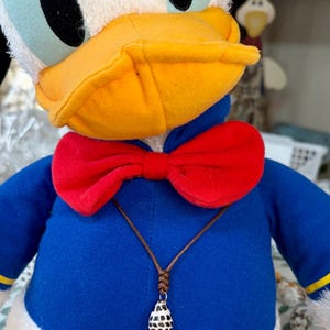 以下が含まれることがあります： 青いシャツに赤い蝶ネクタイをつけたドナルドダックのぬいぐるみ。茶色のコードネックレスには、黒い斑点のある小さな白い涙型のペンダントが付いています。キャラクターは白い頭、黄色いクチバシ、青い目をしています。