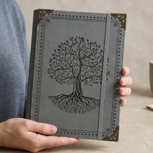 May include: A gray hardcover journal featuring a black embossed tree of life design on the front cover. The notebook includes decorative bronze-toned metal corner protectors and a vertical elastic band closure to keep the pages securely together.