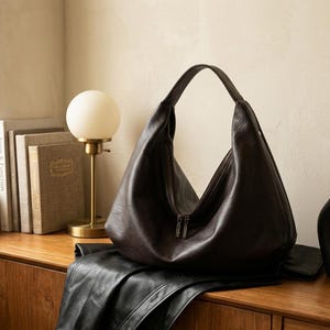 Op de afbeelding: Een donkerbruine leren hobo-tas met ritssluiting, rustend op een houten oppervlak. Een zwarte leren jas ligt onder de tas. Een gouden lamp met een witte bolvormige kap en boeken staan links.