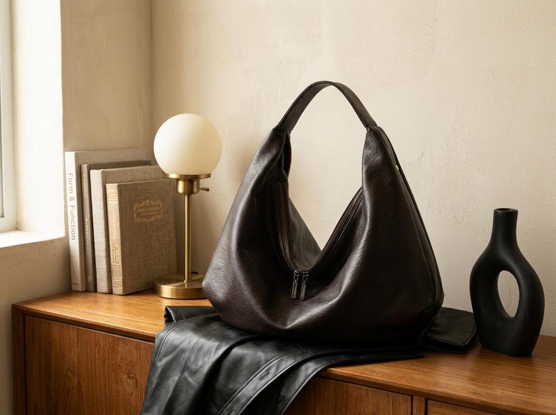 Op de afbeelding: Een donkerbruine leren hobo-tas met ritssluiting, rustend op een houten oppervlak. Een zwarte leren jas ligt onder de tas. Een gouden lamp met een witte bolvormige kap en boeken staan links.