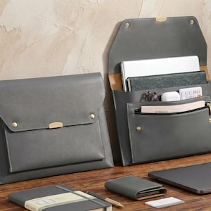 Puede incluir: Dos organizadores de documentos de cuero gris con herrajes dorados. Uno está cerrado, el otro abierto, revelando una computadora portátil, un cuaderno y accesorios. Una cartera, un cuaderno y una computadora portátil a juego también están sobre la superficie de madera.