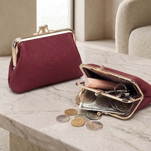 May include: Two burgundy coin purses with gold-coloured frames are on a marble surface. One is closed, the other open, showing coins, keys, and lipstick. The purses have a textured appearance.
