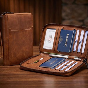 May include: A brown leather travel wallet with a zipper closure, wrist strap, and multiple compartments. The open wallet displays a passport, credit cards, and a pen. The wallet is designed to hold travel documents and essentials.