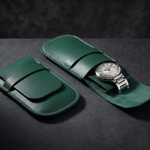 Könnte beinhalten: Zwei grüne Leder-Uhrenetuis. Eines ist offen und zeigt eine silberne Armbanduhr mit weißem Zifferblatt. Das andere ist geschlossen. Beide Etuis haben weiße Nähte und einen Riemenverschluss.