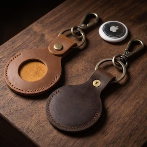May include: Two leather AirTag keychains in brown and dark brown, with metal clasps and key rings. One keychain has a circular cutout, and the other has a brass-colored button. A white Apple AirTag is also visible.
