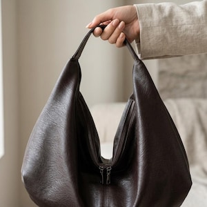 Op de afbeelding: Een donkerbruine hobo-tas met een ritssluiting. De tas is gemaakt van een textuurmateriaal en heeft een enkele handgreep. De tas wordt door een hand omhoog gehouden.